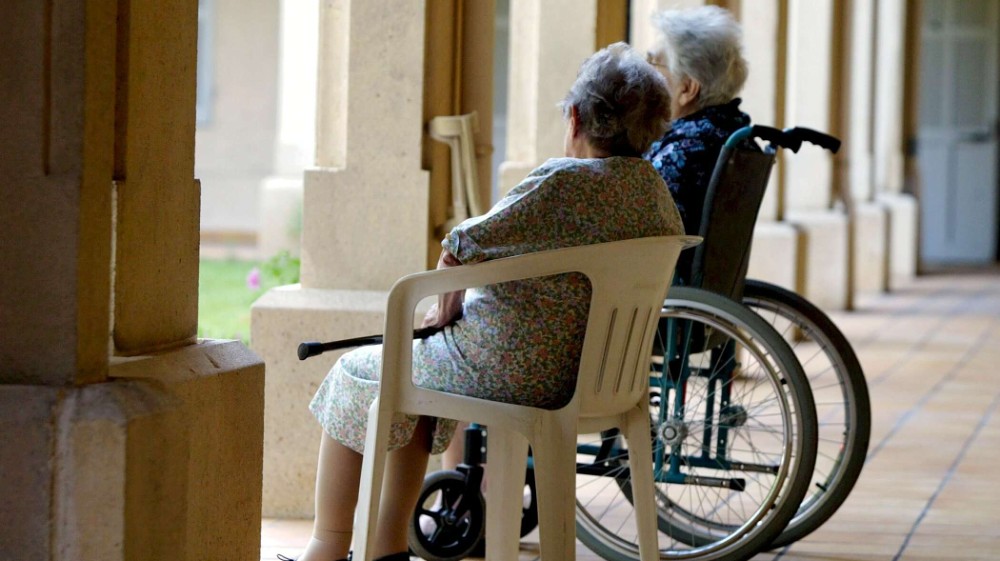 To eldre personer. Den ene sitter i rullestol. Begge ser ut av vinduet. Vi tilbyr privat hjemmehjelp og avlastning.