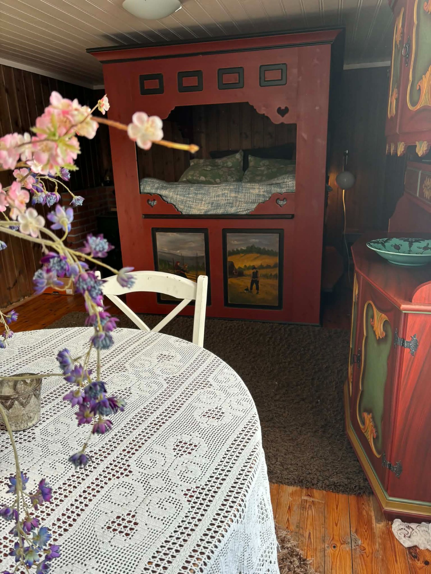 Eldhuset soverom – den rødmalte himmelseng sett forfra, med rosemalt møblement og rundt bord