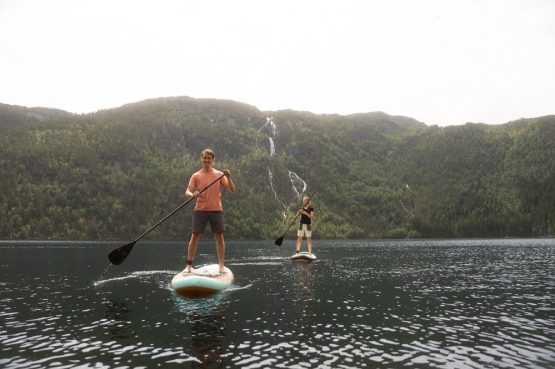 To personer padler SUP på Åkrafjorden, med fossefall i fjellsiden bak