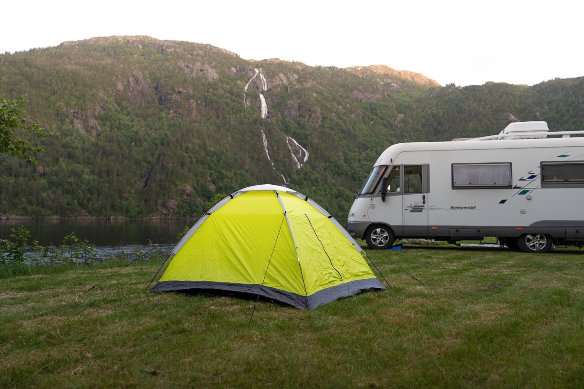 Telt og bobil på gressplenen, med fossefall i fjellsiden bak