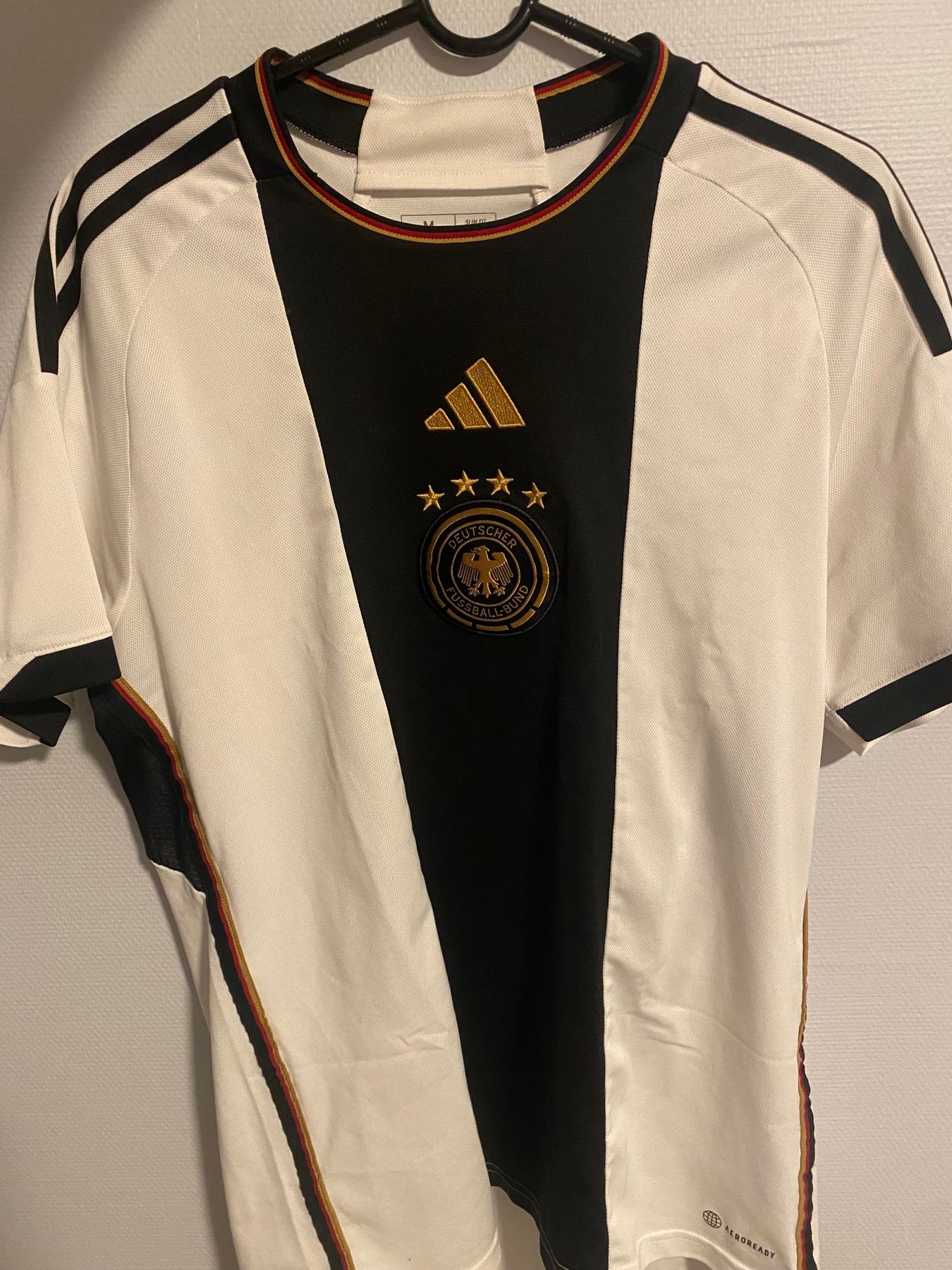 Hvit tysk landslagsdrakt fra VM 2022 sett forfra med bredt svart felt og fire stjerner over logoen