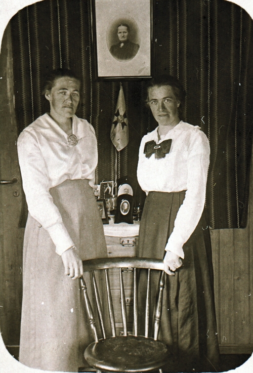 REF-0464 Rennebu ca 1915.
Kari og Ingeborg Flatmo med en stol framfor seg og bilde av mora Karen på veggen. Kari (1886-1966) jordmor i Rennebu. Ingeborg (1889-1983) lærar bl.a.i Tydal og på Refshus.