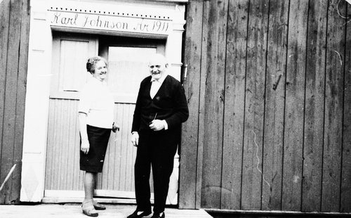 REF-0511 Stølen 1969. Ane Johnson f. Brattset og Karl Johnson framfor inngangsdøra.Over døra står malt 'Karl Johnson År 1911'. Karl(1891-1978) bureisar i Stølen, ordførar i Rennebu. Ane (1896-1978) bureisarkone, formann i sanitetsforeninga mm.